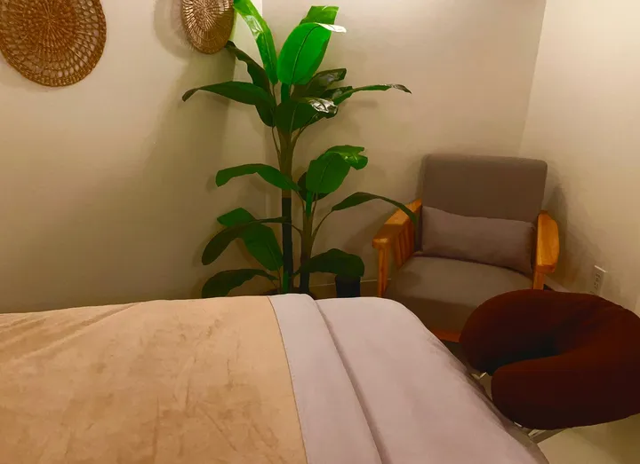 A massage table in a calm room with a tall plant and a soft chair