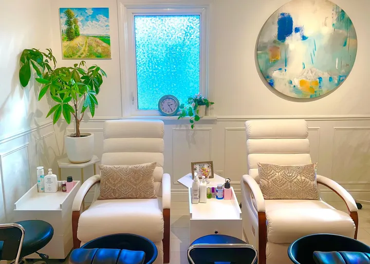Two white pedicure chairs under a bright window, with plants and paintings